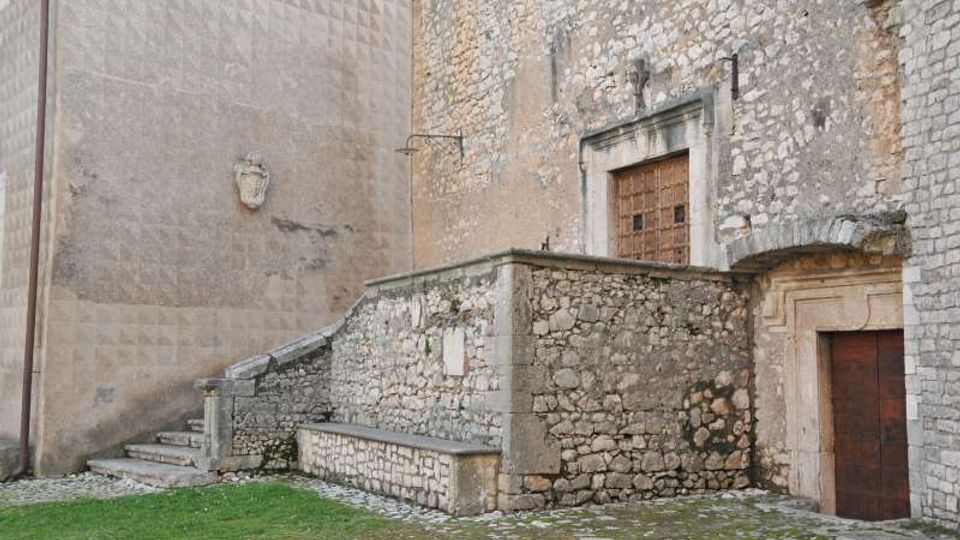 Cortile in pietra con scala laterale e muretto in muratura, addossati a una parete di edificio in pietra e intonaco decorato. Si vedono due porte in legno con cornici in pietra e un piccolo stemma sulla parete.