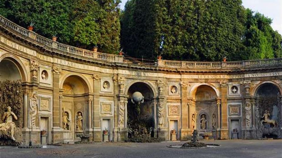 Esedra monumentale in pietra con grandi arcate e nicchie ornate da statue e rilievi, disposta a semicerchio attorno a una piazza pavimentata. Sullo sfondo si vedono alberi alti e una balaustra superiore che corre lungo la sommità della struttura.