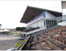 Tribuna di uno stadio o ippodromo con gradinate in cemento e file di sedili in plastica marrone, affiancata da un edificio con ampie vetrate e grande tetto sporgente. In basso si vedono l’area esterna con strada, parcheggio e alcune strutture temporanee.