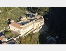 Veduta aerea di un grande complesso monastico in pietra chiara con edifici a tetto in coppi e una torre campanaria, disposto attorno a cortili interni. La struttura è costruita sul bordo di un pendio boscoso, con oliveti e strade di accesso nelle vicinanze.