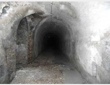 Tunnel sotterraneo stretto con volta a botte in cemento grezzo, pareti macchiate e tratti di mattoni a vista sul lato sinistro. Il pavimento è parzialmente coperto da detriti e il corridoio si estende in profondità verso un’area più buia.