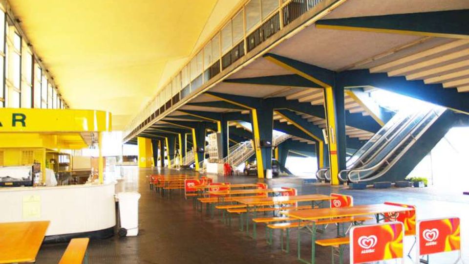 Ampio spazio interno tipo atrio o area ristoro con tavoli e panche in fila, pavimento scuro e chiosco bar laterale. Struttura industriale con colonne e travi in acciaio verniciate, grandi finestre e scale mobili che salgono al piano superiore.