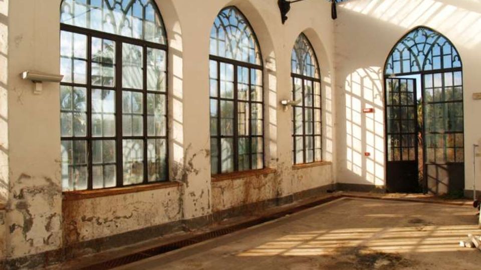 Interno di una serra o padiglione con alte finestre ad arco in ferro e vetro lungo le pareti e una copertura parzialmente vetrata. Pavimento in cemento con canaletta di drenaggio, pareti intonacate segnate dall’usura e una porta vetrata sul fondo.
