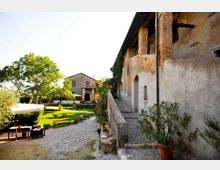 Cortile di un casale rustico in pietra e intonaco, con lungo portico ad archi e camminamento rialzato lungo la facciata. In primo piano un vialetto in ciottoli conduce a un’area verde con alberi, tavoli e panche all’aperto, e un edificio secondario sullo sfondo.