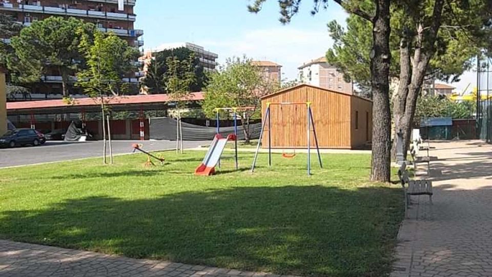 Piccolo parco urbano con prato e area giochi con altalene e scivolo, affiancata da un vialetto pavimentato e panchine sotto alti pini. Sullo sfondo si vedono una casetta in legno e palazzi residenziali.