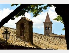 Tetti in coppi di un borgo storico con un piccolo campanile in pietra e, sullo sfondo, una torre campanaria con cuspide. L’inquadratura è incorniciata da rami e foglie di albero, con un lampione in ferro in primo piano.