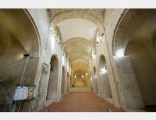 Interno di una chiesa o cripta in pietra con lunga navata centrale, alte arcate laterali e volte a botte illuminate da faretti. In fondo si vede una breve scalinata che conduce all’area dell’altare.