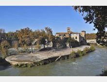Isola fluviale con una piazza pavimentata e gradinate semicircolari affacciate sull’acqua, circondata da alberi. Sullo sfondo si vedono edifici storici con una torre e un ponte ad arco che attraversa il fiume.