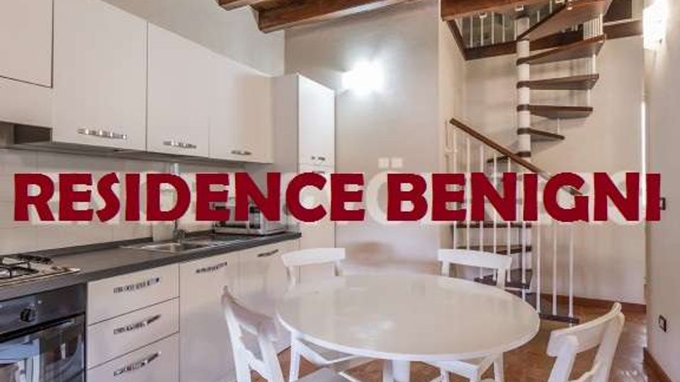 Cucina-soggiorno open space con mobili bianchi e piano cottura lineare lungo la parete, tavolo rotondo con sedie al centro e soffitto con travi in legno a vista. Sul lato destro si vede una scala a chiocciola in metallo che conduce al piano superiore.