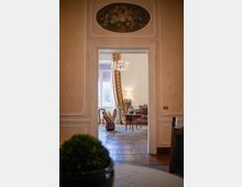 Interno di un appartamento elegante con pareti in stucco e cornici decorative, con un arco sopra la porta che incornicia un dipinto ovale di fiori. Attraverso la porta si vede un salotto con lampadario, grandi finestre con tende pesanti, parquet a spina e mobili classici con poltrone a righe e tavolino.