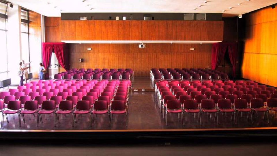 Sala conferenze con pareti rivestite in legno, soffitto bianco con illuminazione a incasso e file di sedie imbottite bordeaux disposte in blocchi con corridoio centrale. Sul lato sinistro grandi finestre a tutta altezza e tende bordeaux incorniciano l’area frontale della sala.