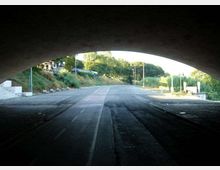 Strada asfaltata vista dall’interno di un sottopasso ad arco, con corsia ciclabile centrale e carreggiata che prosegue in leggera curva. Ai lati si vedono scarpate erbose, alberi e alcuni lampioni lungo il margine della strada.