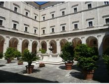 Cortile interno di un edificio in pietra con portico ad archi su più lati e facciate con numerose finestre rettangolari. Al centro si trova una fontana monumentale, circondata da vasi con alberi e piante su pavimentazione in pietra.