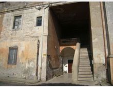 Facciata di un edificio antico con intonaco consumato e finestre rettangolari, affacciata su una strada. A destra si apre un grande passaggio coperto con una scalinata in pietra che sale lungo il muro e conduce a un cortile interno con arco e portone.