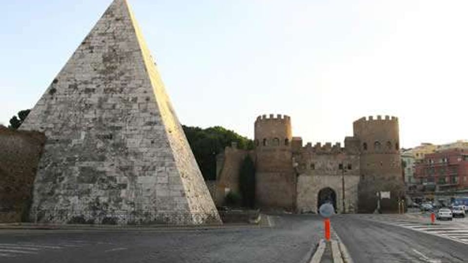 Piazza con ampia piramide rivestita in pietra in primo piano e, sullo sfondo, una fortificazione in mattoni con mura merlate, due torri e un grande arco d’ingresso. Strada asfaltata e area urbana circostante visibili ai lati.