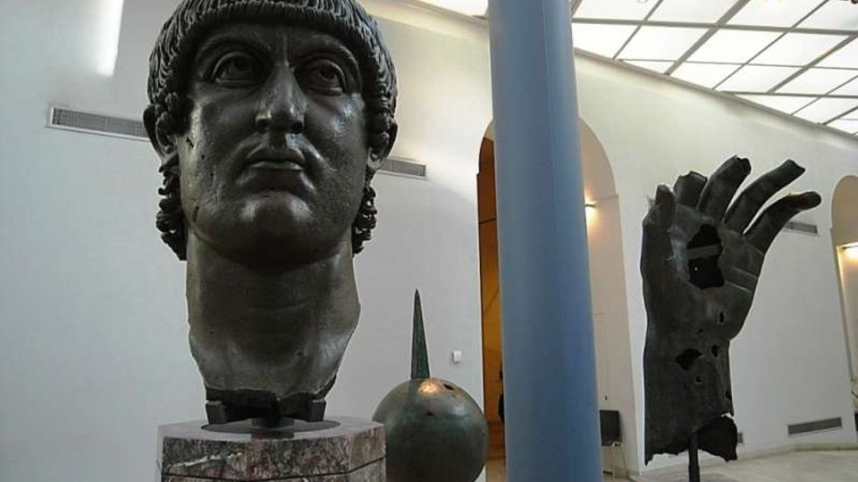 Sala museale luminosa con pareti bianche, lucernario sul soffitto e colonne, con grandi sculture scure su piedistalli in marmo, tra cui una testa monumentale e una mano. Sullo sfondo si vedono archi e altre opere esposte nello spazio.