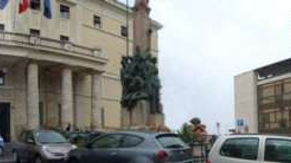 Veduta panoramica di una piazza urbana con parcheggio in primo piano, affacciata su un edificio istituzionale con portico a colonne e bandiere e su palazzi residenziali moderni a più piani. Sullo sfondo si vedono altre costruzioni e il cielo nuvoloso.