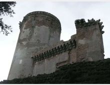 Vista dal basso di una fortificazione in muratura con una torre cilindrica e un mastio rettangolare, entrambi con merlature e beccatelli sporgenti. Le pareti mostrano intonaco consumato e mattoni a vista, sopra una scarpata rocciosa con rami d’albero ai margini del cielo.