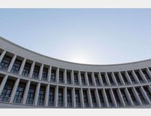 Facciata curva di un grande edificio moderno con file regolari di colonne verticali e finestre su più piani, ripresa dal basso verso l’alto. Sullo sfondo si vede il cielo limpido.