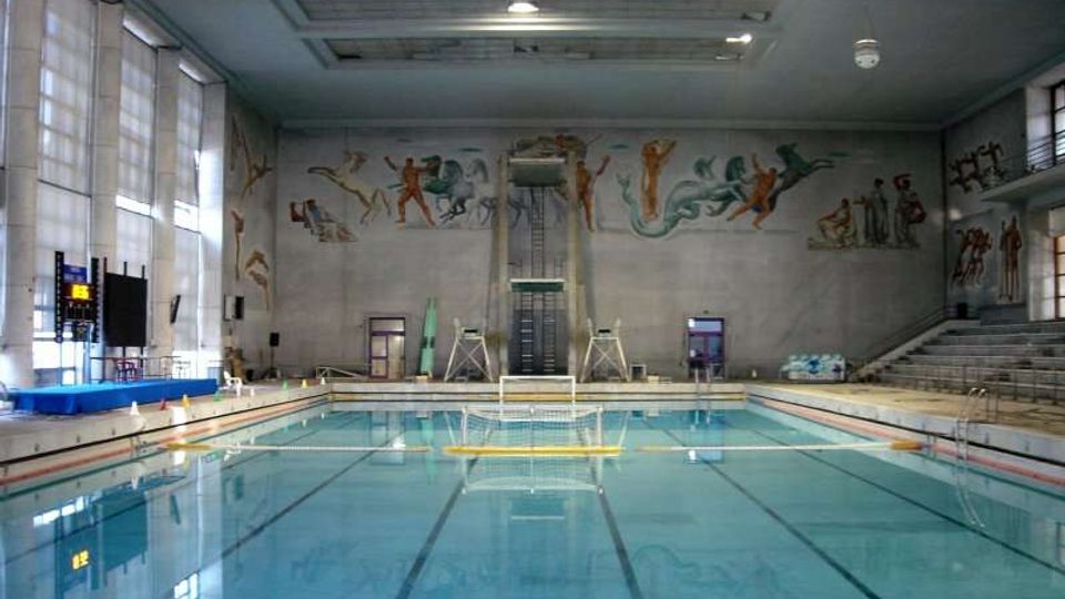 Piscina coperta con grande vasca rettangolare e corsie, illuminata da ampie finestre laterali. Sul fondo si vede una piattaforma per tuffi e grandi murales figurativi sulle pareti, con gradinate per spettatori su un lato.