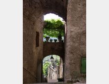 Passaggio stretto in pietra con grandi archi sovrapposti, che incorniciano una terrazza con ringhiera e piante in vaso sotto una pergola verde. In fondo si vede una discesa tra muri antichi con un lampione sospeso e vegetazione sullo sfondo.