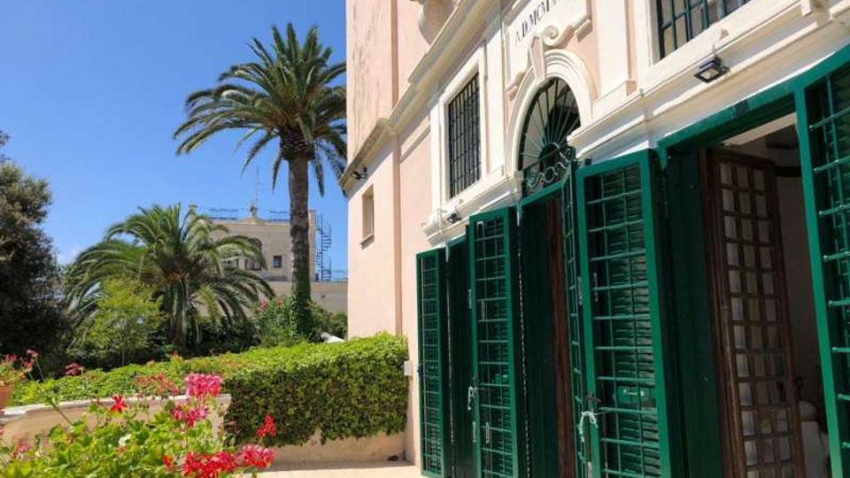 Facciata di un edificio chiaro con ingresso ad arco e grandi persiane verdi aperte, affacciato su un cortile pavimentato. Accanto all’edificio si vede un giardino con siepi, fiori e alte palme sotto un cielo sereno.