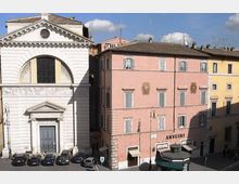 Facciate su una piazza urbana: a sinistra un edificio bianco in stile neoclassico con grande frontone e portale, a destra un palazzo rosa a più piani con persiane e insegna “ANSUINI”. In basso si vedono auto parcheggiate lungo il bordo della piazza e lampioni stradali.