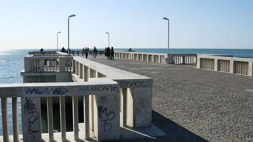 Molo sul mare con ampia pavimentazione in pietra e parapetti in cemento segnati da graffiti, che si estende verso l’orizzonte. Lungo il percorso sono presenti alti lampioni e si vede il mare aperto su entrambi i lati.
