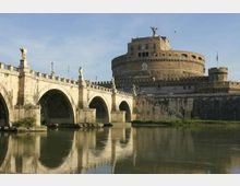 Veduta di Castel Sant’Angelo a Roma con il Ponte Sant’Angelo in primo piano, composto da arcate in pietra e statue lungo il parapetto. Il fiume Tevere scorre sotto il ponte e riflette la fortezza cilindrica con bastioni e mura.