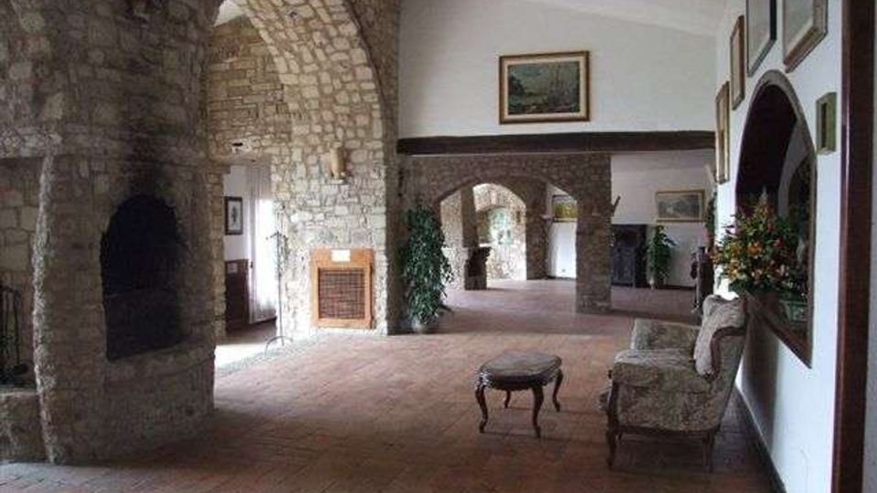 Ampio salone interno con pavimento in cotto e pareti in pietra e intonaco, attraversato da grandi archi in muratura che conducono a corridoi e stanze sullo sfondo. Sulla destra sono presenti specchi e quadri alle pareti e un piccolo salottino con poltrone e tavolino, con piante e decorazioni floreali.