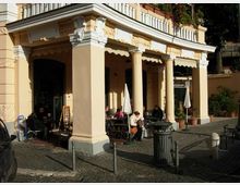 Portico di un edificio storico color crema con grandi colonne e decorazioni in stucco, affacciato su una piazzetta con pavimentazione in sampietrini. Sotto il portico si vede l’area esterna di un bar con tavolini, sedie e ombrelloni chiusi, circondata da piante in vaso e paletti lungo il marciapiede.