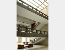 Atrio interno di un edificio moderno a più livelli, con passerelle e rampe in pietra scura e parapetti in vetro. Un lucernario a griglia sul soffitto illumina lo spazio con grandi superfici vetrate e un’area centrale con panchine.