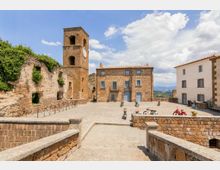 Piazza lastricata in un borgo storico con edifici in pietra e una torre campanaria quadrata con orologio; a sinistra si vedono muri diroccati coperti di vegetazione. Sullo sfondo compaiono case con tetti in coppi e un panorama collinare sotto un cielo parzialmente nuvoloso.