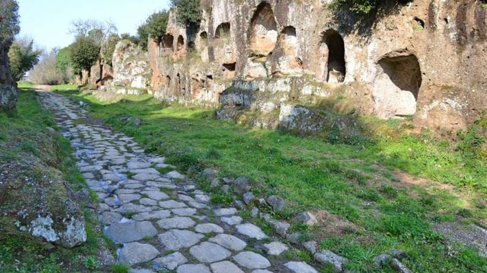 Sentiero in pietra irregolare che attraversa un’area erbosa, costeggiando una lunga parete di roccia tufacea con numerose cavità e nicchie scavate. Vegetazione bassa e alberi crescono sopra la scarpata rocciosa lungo il percorso.