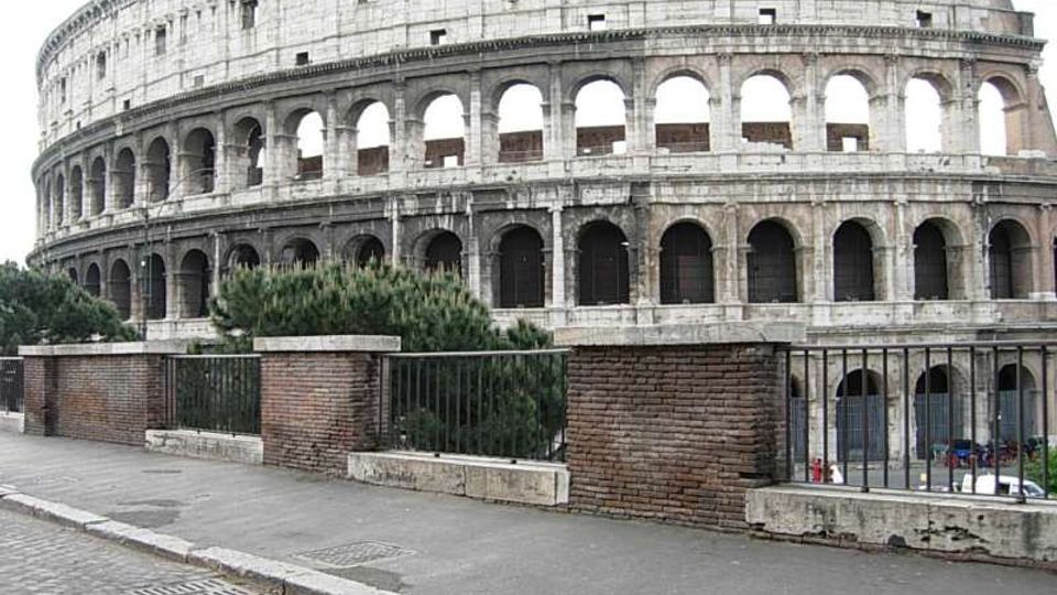 Veduta esterna del Colosseo, anfiteatro in pietra con più ordini di arcate sovrapposte e ampie aperture. In primo piano si vede un marciapiede con muretto in mattoni e ringhiera metallica, con cespugli lungo il bordo.