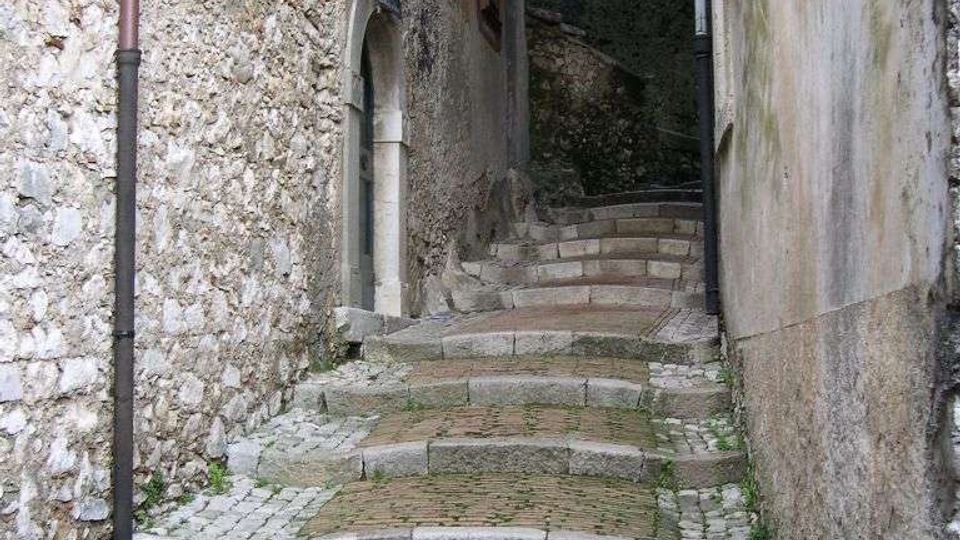 Vicolo stretto in pietra con pavimentazione a ciottoli e gradini in mattoni e lastre, tra muri di edifici antichi. In fondo si vede un passaggio ad arco coperto con una lanterna e una breve rampa che sale.