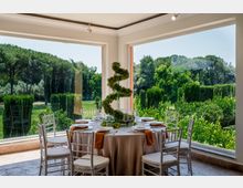 Sala da pranzo luminosa con ampie finestre panoramiche ad angolo, tavolo rotondo apparecchiato e sedie bianche. All’esterno si vede un grande giardino verde con siepi e alberi alti.