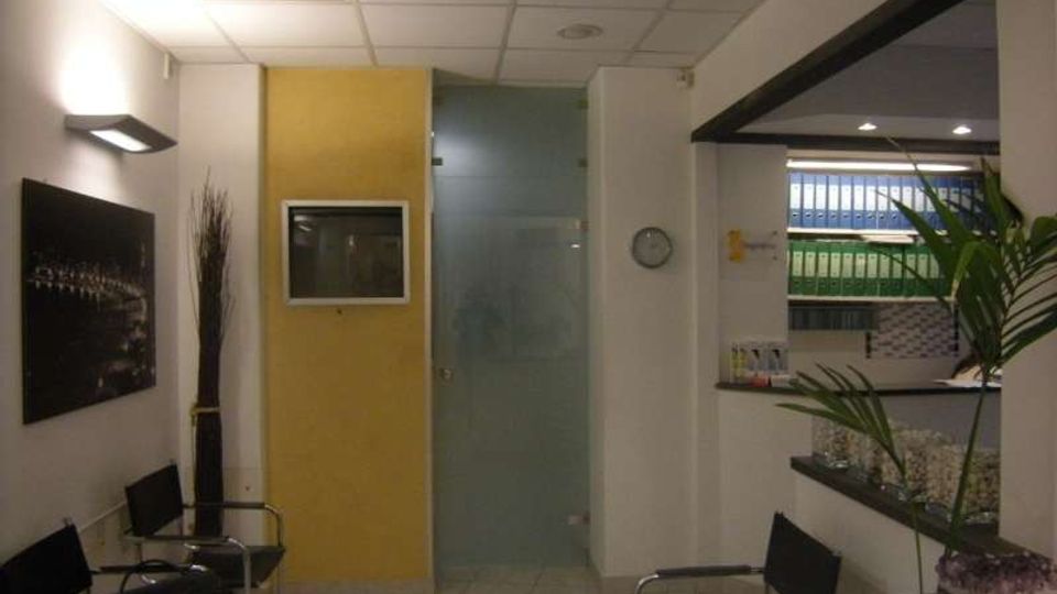 Interno di una sala d’attesa in ufficio, con sedie nere lungo la parete, pavimento piastrellato e controsoffitto a pannelli con luci. Si vedono una parete gialla con display incassato, una porta in vetro satinato, un bancone con scaffali di raccoglitori e alcune piante decorative.