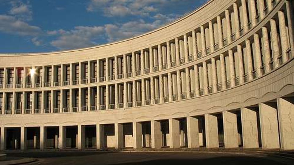 Edificio monumentale a pianta curva con lunga facciata in pietra, scandita da file regolari di alte finestre e colonne. In primo piano si vede un porticato ad archi che si affaccia su un’ampia area pavimentata, sotto un cielo parzialmente nuvoloso.