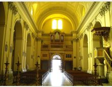Interno di una chiesa con navata centrale lunga e simmetrica, volta a botte e arcate laterali su colonne. In fondo si vedono la cantoria con grande organo a canne, la porta d’ingresso luminosa e, sulla destra, un pulpito decorato.