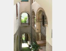 Vicolo stretto tra edifici in muratura chiara con porticati ad archi in pietra e un cancelletto in ferro battuto, con piccoli gradini e piante in vaso. In fondo si vedono altre arcate e uno scorcio di vegetazione e colline sullo sfondo.