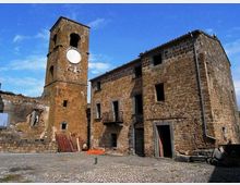 Piazza lastricata in pietra con edifici rurali in muratura di tufo, tra cui una torre campanaria con quadrante circolare e un fabbricato a due piani con piccole finestre e portali ad arco. Sullo sfondo cielo azzurro e alcune strutture parzialmente diroccate ai margini della piazza.