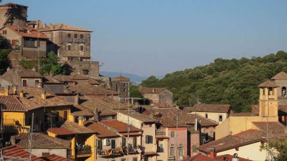 Panorama di un borgo collinare con case addossate dai tetti in coppi e facciate colorate, dominato da una torre cilindrica e da un grande edificio storico in pietra. A destra si vede un campanile e, sullo sfondo, un’area boscosa con colline sotto un cielo sereno.
