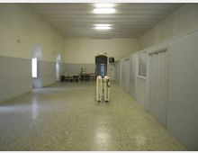 Lungo corridoio interno con pavimento in graniglia e pareti bicolori, illuminato da plafoniere a soffitto. Sul lato destro si vedono pareti divisorie con porte e finestre, mentre in fondo ci sono alcune sedie e tavoli vicino a una porta ad arco.