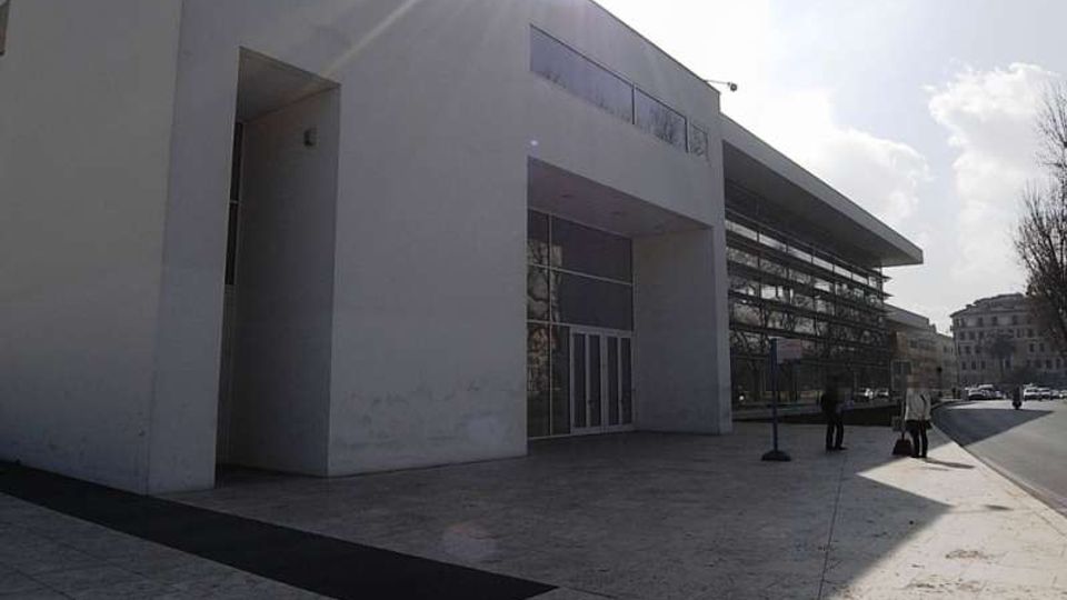 Facciata di un edificio moderno con grandi volumi bianchi e ingresso incassato con porte a vetri, affacciato su un ampio piazzale pavimentato. Sul lato destro si vede una strada urbana con marciapiede, alberi e altri edifici sullo sfondo.