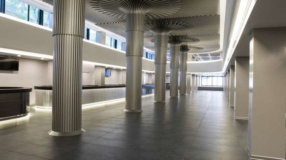 Ampio atrio interno di un edificio moderno con pavimento in grandi piastrelle scure, illuminazione lineare a soffitto e una fila di colonne metalliche scanalate con capitelli a raggiera. Ai lati si vedono banconi bassi e finestre alte che lasciano entrare luce naturale, con vista verso un’area vetrata in fondo.