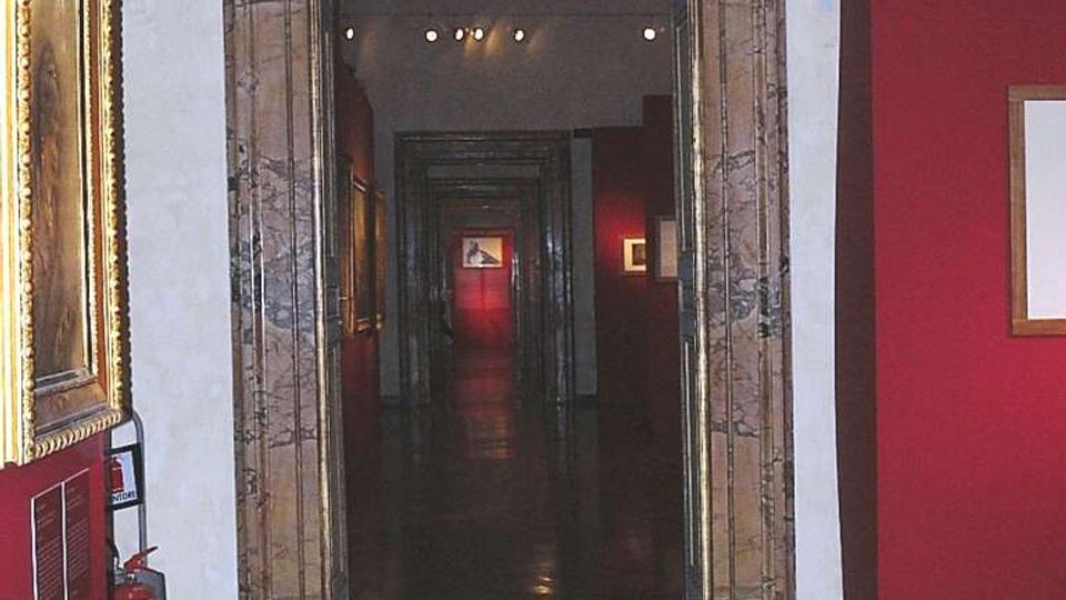 Interno di museo con pareti rosse e pavimento in legno scuro lucido, visto attraverso un grande portale rettangolare con cornice in marmo. In fondo si apre un lungo corridoio in prospettiva con più passaggi allineati e quadri esposti alle pareti, illuminato da luci a soffitto.