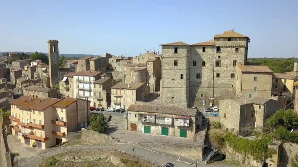 Veduta panoramica di un borgo in pietra con case addossate e una grande struttura fortificata simile a un castello al centro, affiancata da una torre campanaria. In primo piano si vedono una strada e un terrapieno, con colline verdi sullo sfondo sotto un cielo sereno.