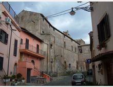 Stradina in pietra in un borgo storico, fiancheggiata da edifici in intonaco rosa e grigio con persiane, balconi e cavi aerei. Sullo sfondo si vede una struttura in muratura dall’aspetto medievale e un lampione sospeso sopra la via.