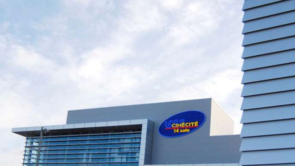 Esterno di un moderno complesso cinematografico con ampia facciata vetrata e rivestimenti metallici, con insegna “Cinecitté 14 sale” su un volume superiore. Davanti si apre una grande area pedonale pavimentata, con un edificio alto che incornicia la scena sul lato destro sotto un cielo nuvoloso.